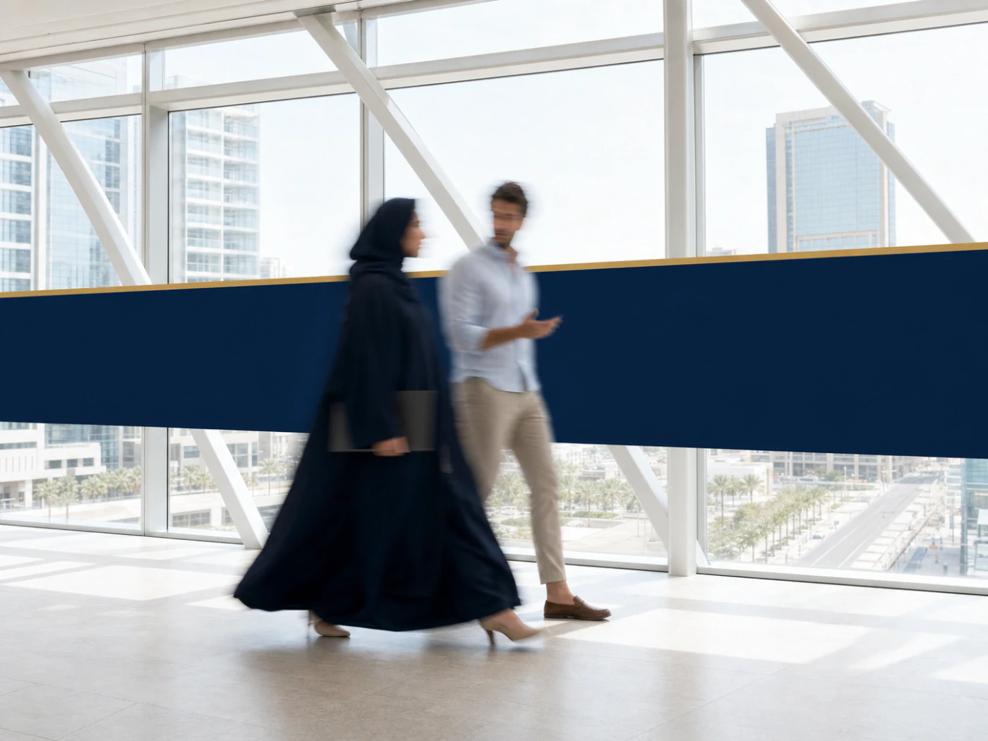 Two motion-blurred professionals crossing a bright glass skybridge.